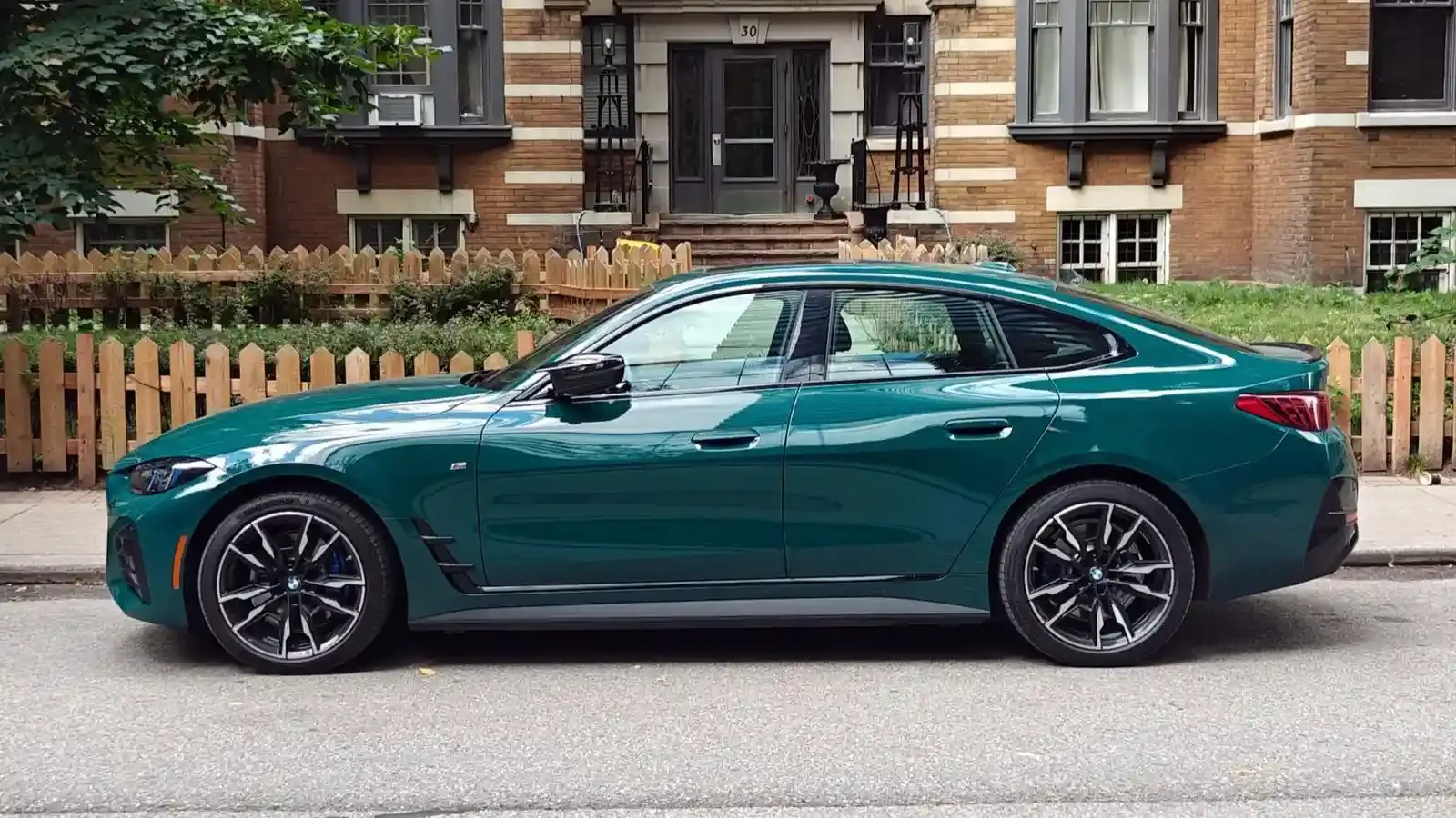 Side profile of a Cape York Green 2026 BMW i4 M60 xDrive parked on the curb in front of a brick building with a picket fence