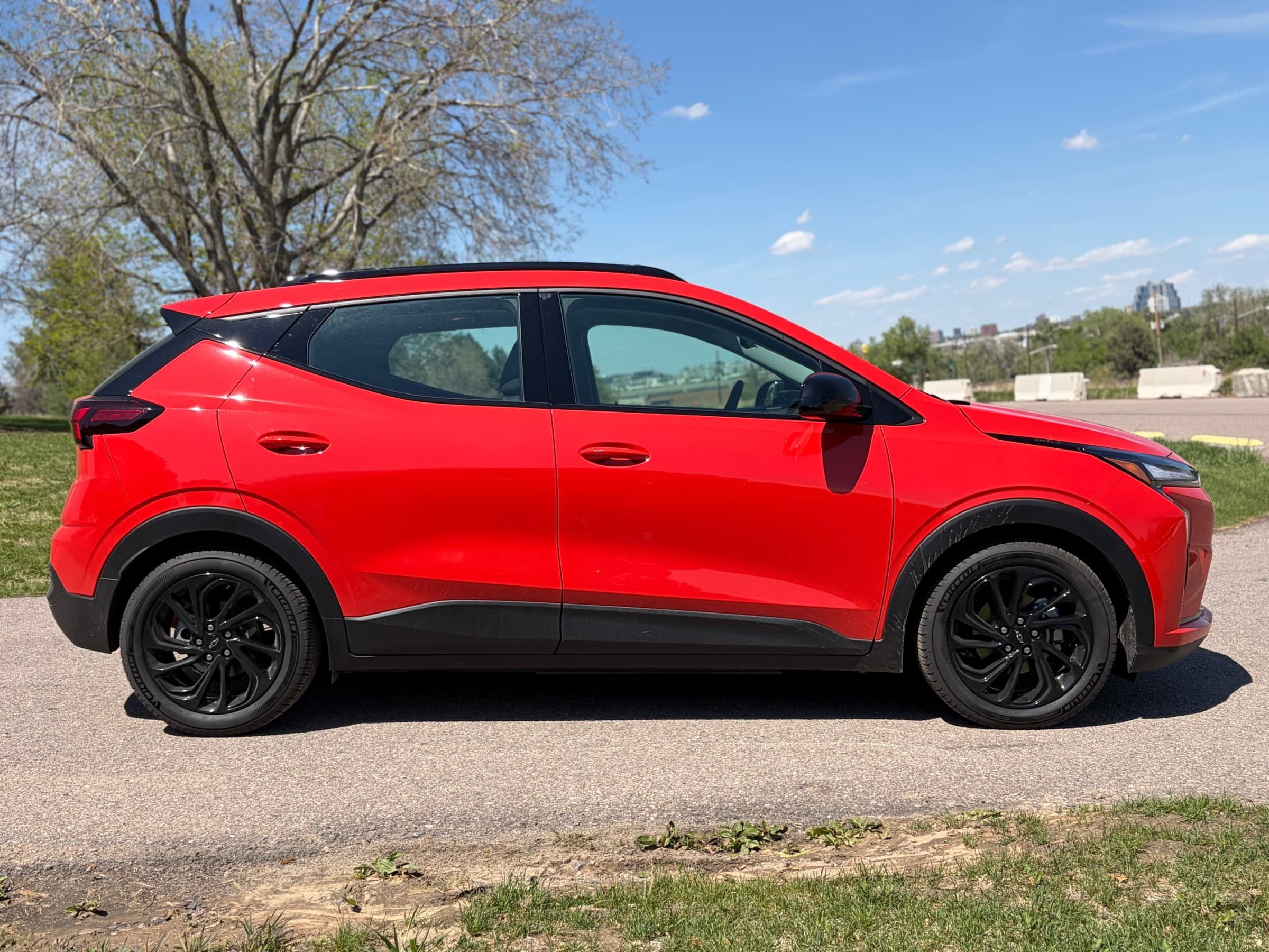 Side profile of a red Chevrolet Bolt with a black roof and black wheels, parked on a path with a city skyline in the distance.