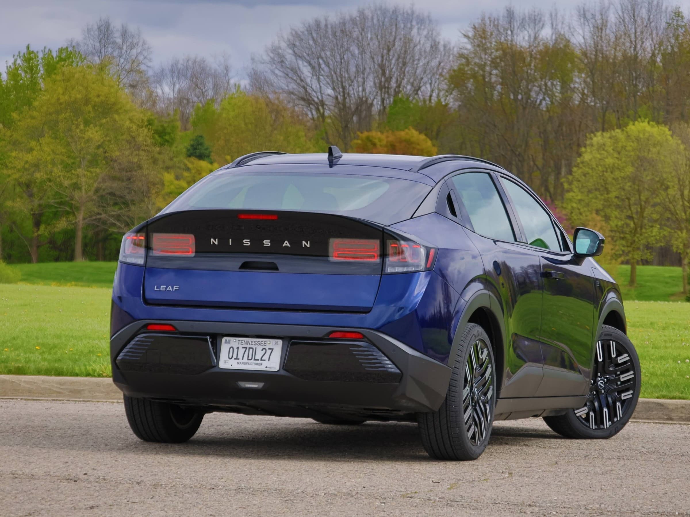  Rear three-quarter view of a dark blue Nissan Leaf parked in front of spring trees, showing its sloping roofline and black wheels.