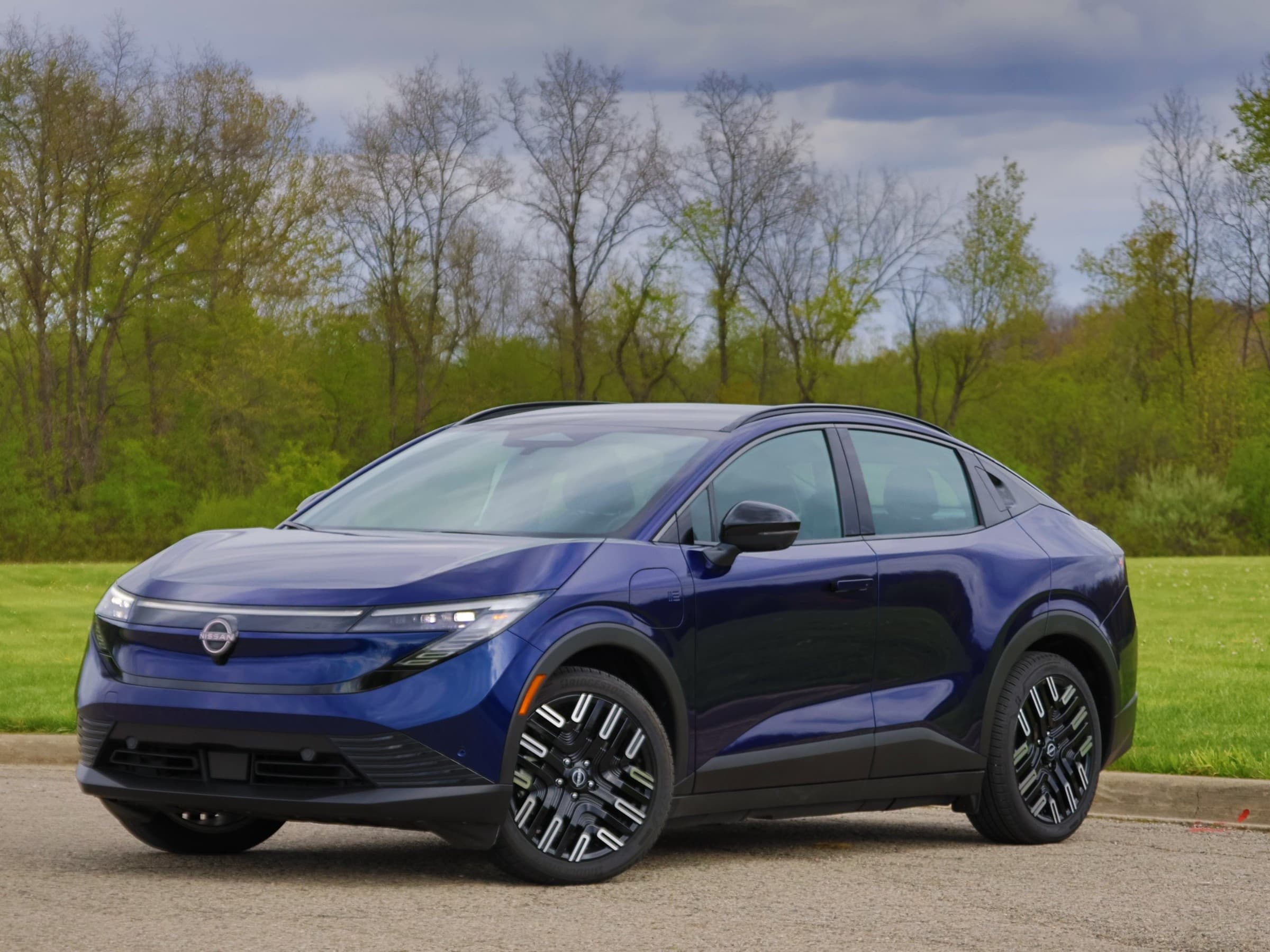 Front three-quarter view of a dark blue Nissan Leaf with a light-bar front end and two-tone wheels, parked beside a grassy field.