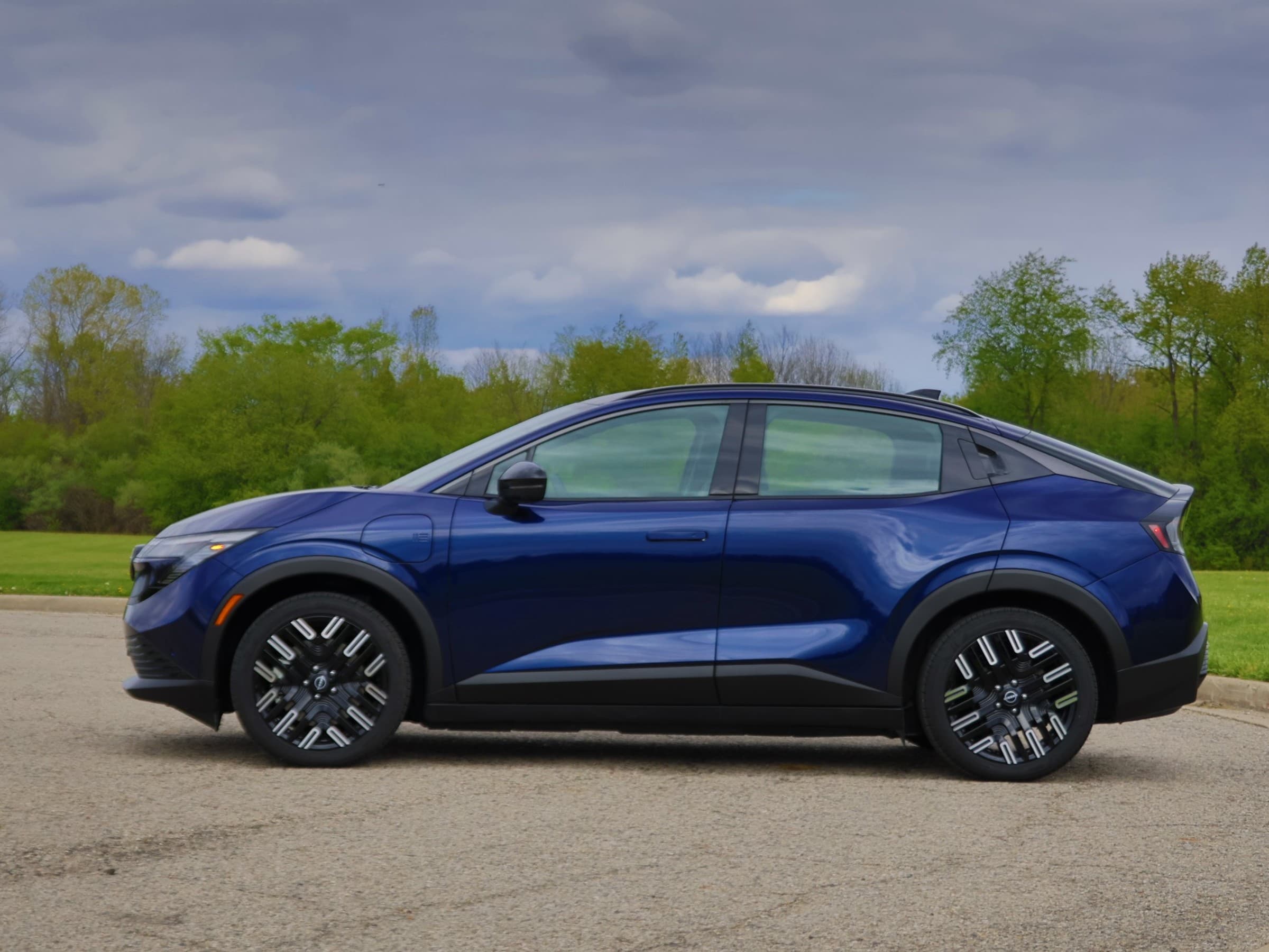 Side profile of a dark blue Nissan Leaf crossover showing its coupe-like roofline and flush door handles, with trees behind.