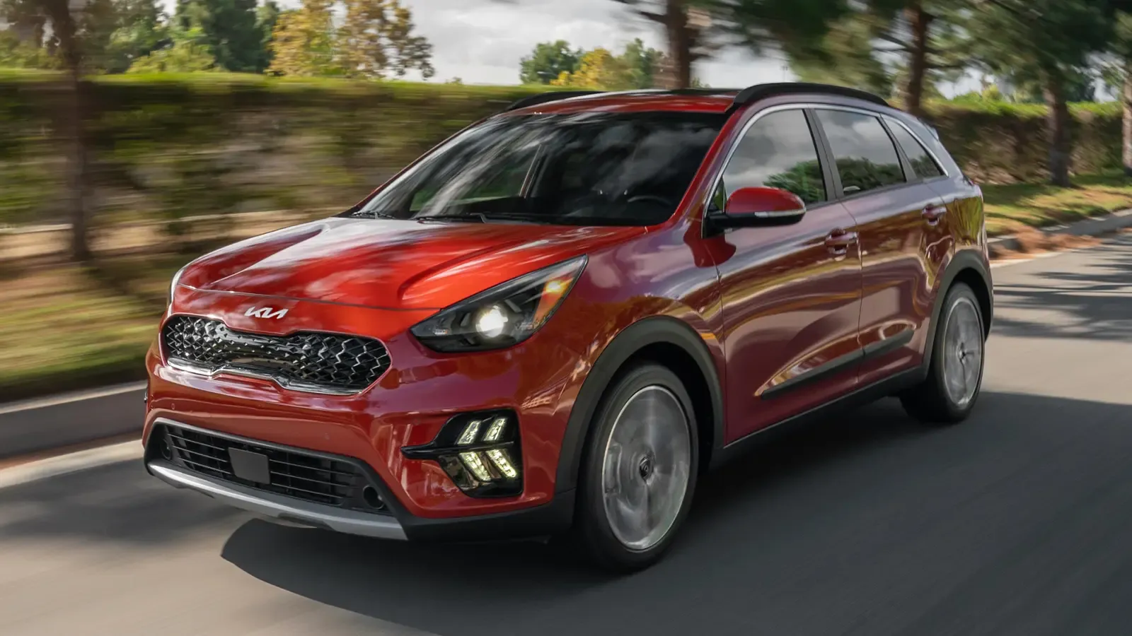kia niro driving down a street with bushes, grass and trees in the background