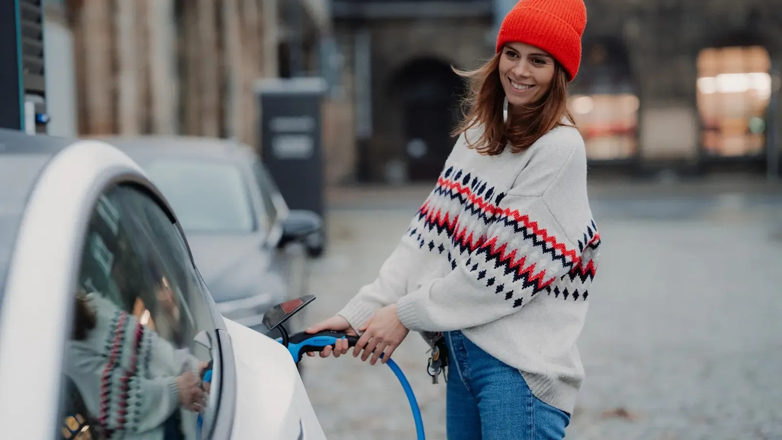smiling woman wearing orange hat and sweater plugging in and charging her electric car
