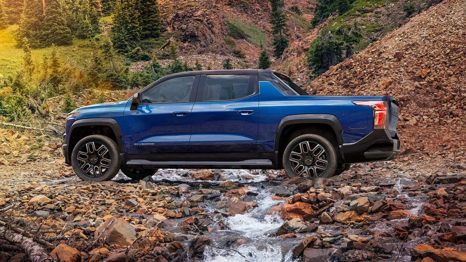 Side view of the Chevrolet Silverado EV driving on a forest path by the mountains