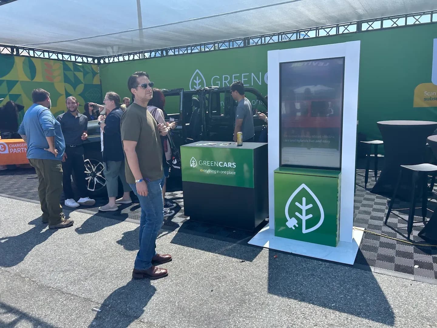 Varias personas se reúnen junto a un stand de GreenCars al lado de un vehículo todoterreno negro.