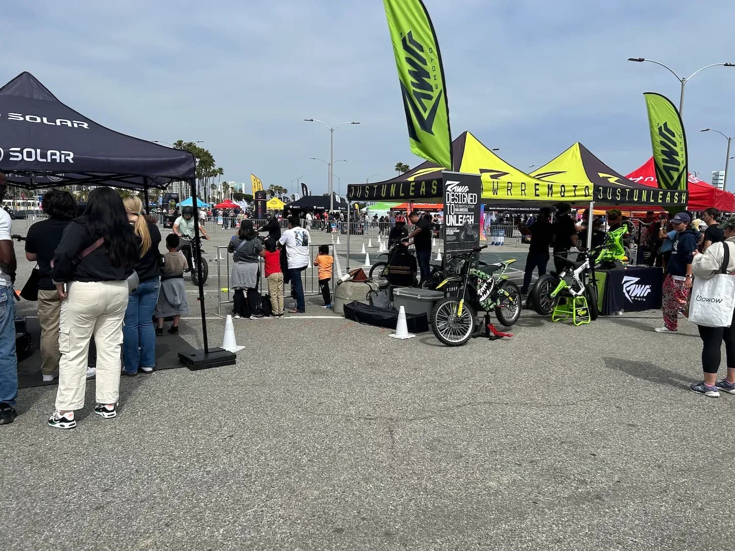 Vista amplia de una expo al aire libre con personas reunidas alrededor de exhibiciones de bicicletas eléctricas bajo carpas con marca.