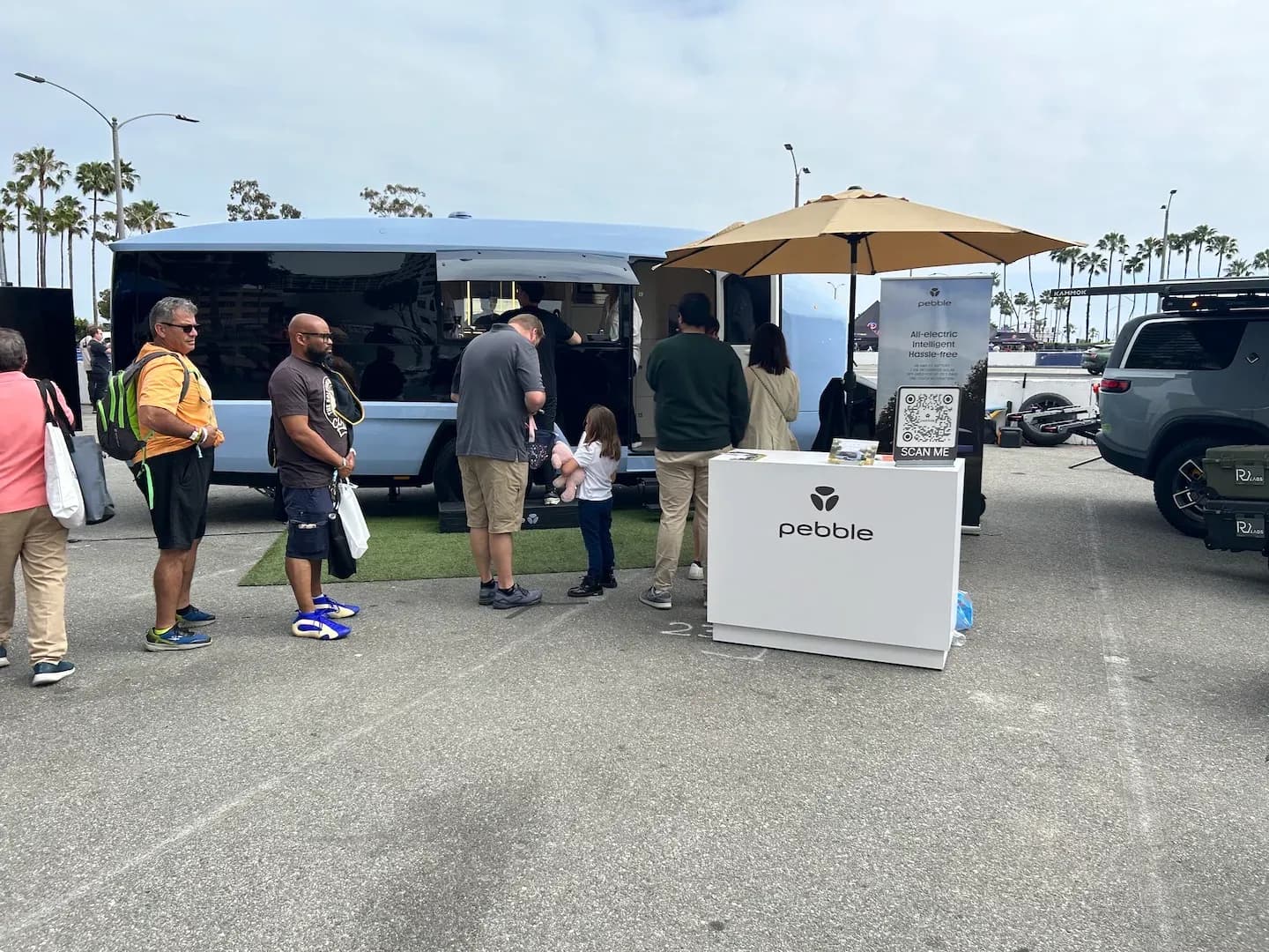 Vista lateral de un remolque camper eléctrico azul con varias personas reunidas alrededor de un stand de Pebble.