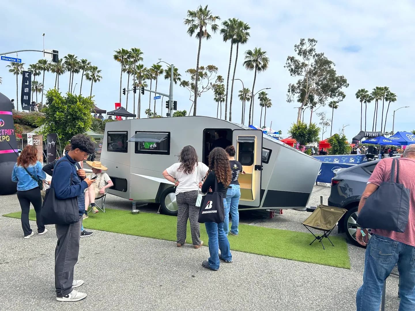 Vista lateral de una caravana gris futurista con varias personas reunidas alrededor en una expo al aire libre.
