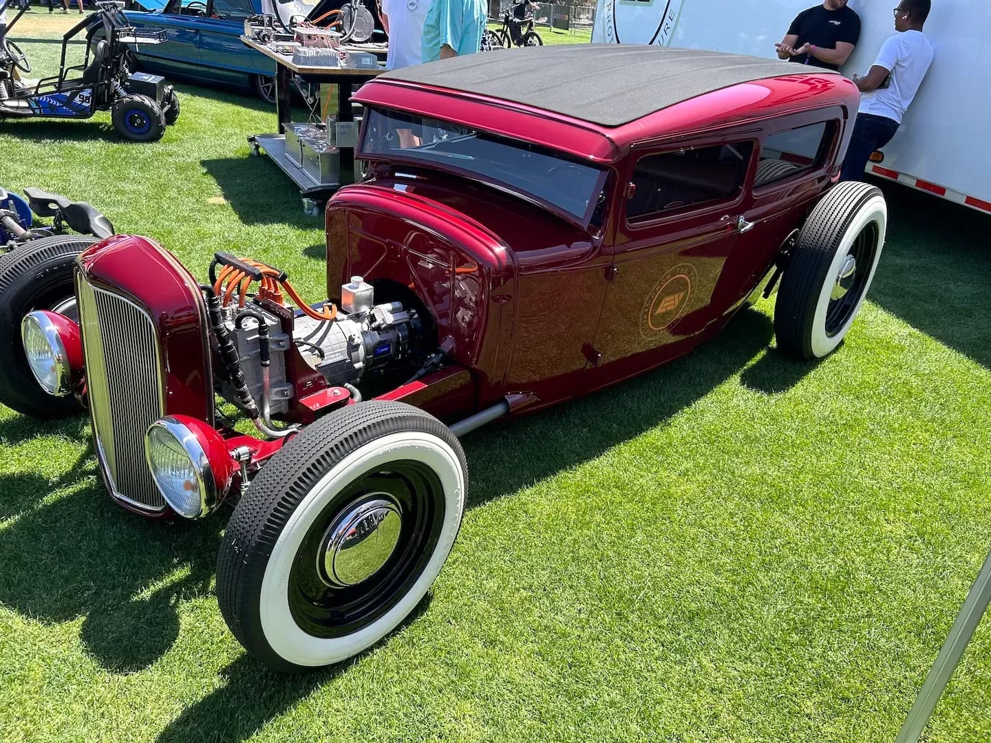 Vista frontal de tres cuartos de un hot rod personalizado color vino con tren motriz eléctrico expuesto en una expo al aire libre.