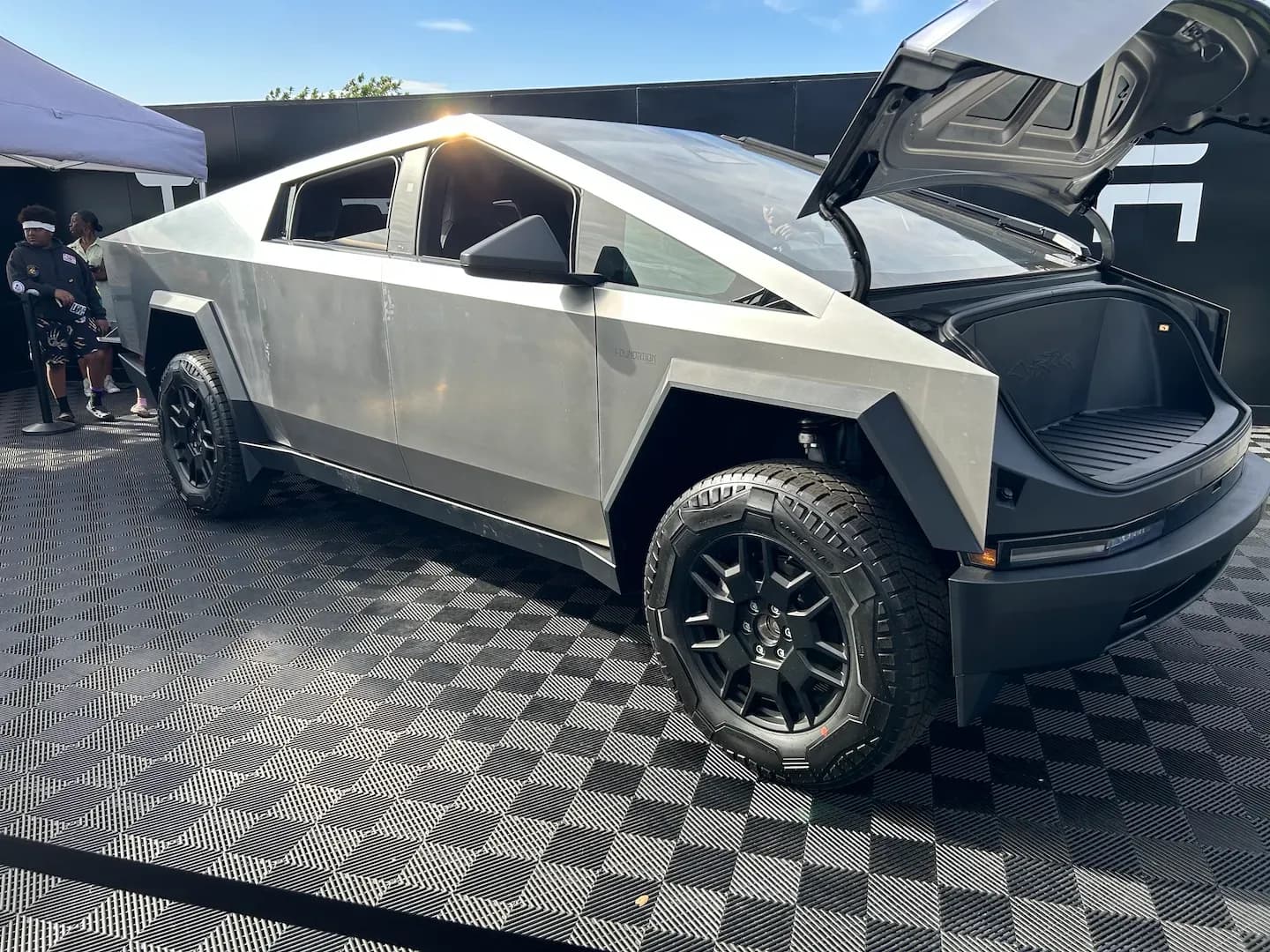 Vista trasera de tres cuartos de una pickup eléctrica plateada con el cofre delantero abierto en una expo.