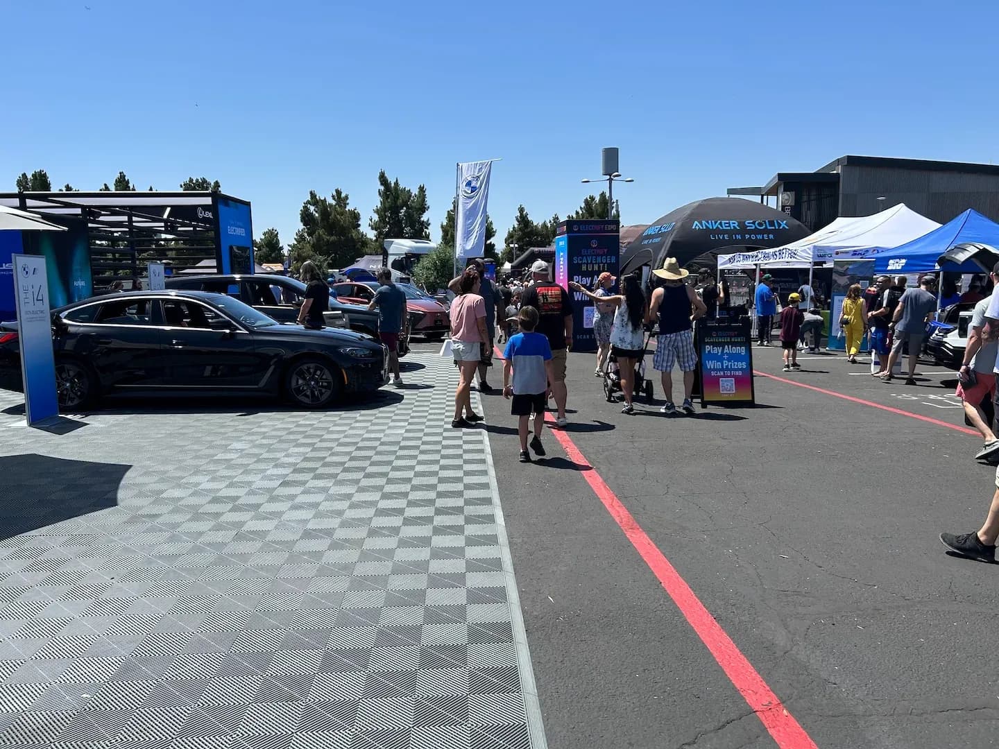 Plano general de personas caminando entre exhibiciones de autos eléctricos y carpas de vendedores en una expo al aire libre.