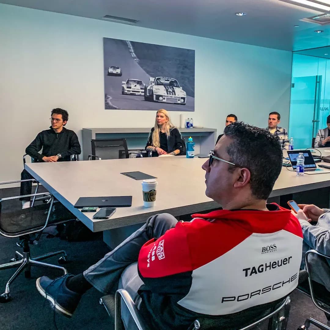 Personas sentadas alrededor de una mesa de conferencia en una sala de reuniones con un póster de carreras de Porsche en la pared.