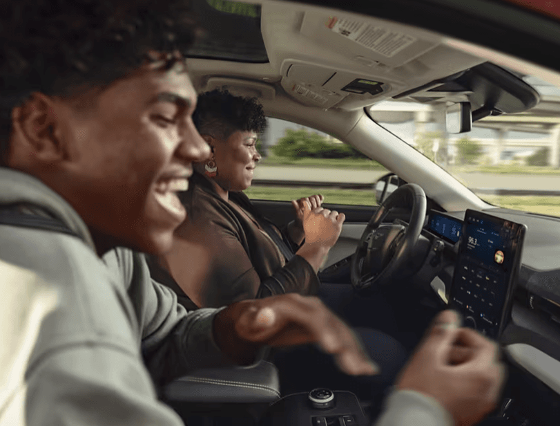 Interior side view of two people in the front seats of a car, with the driver’s hands on the wheel.