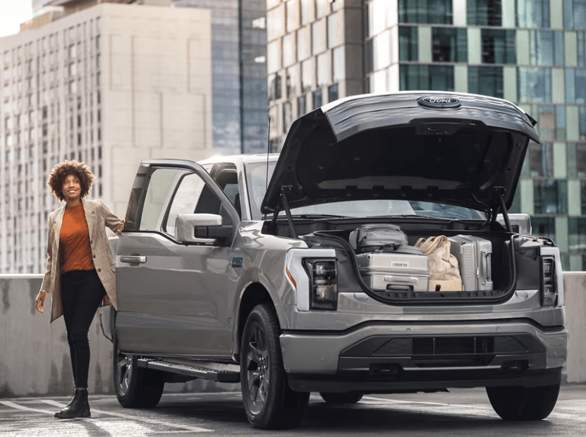 A person stands beside a silver electric pickup with the front trunk open and luggage inside.