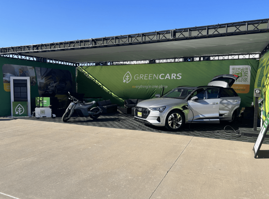 Una SUV eléctrica plateada está conectada junto a una motocicleta en el puesto de GreenCars en la expo.
