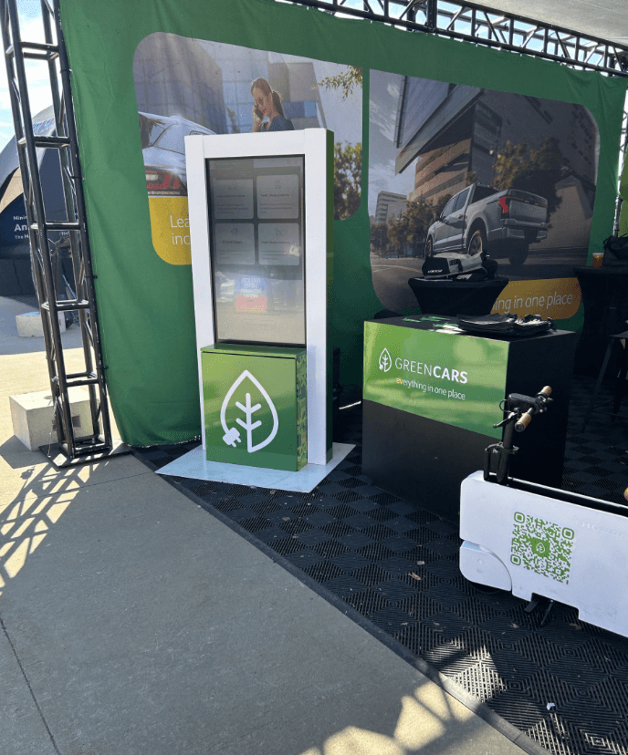 Stand de GreenCars con un quiosco táctil, una mesa con la marca y un patinete eléctrico en exhibición.