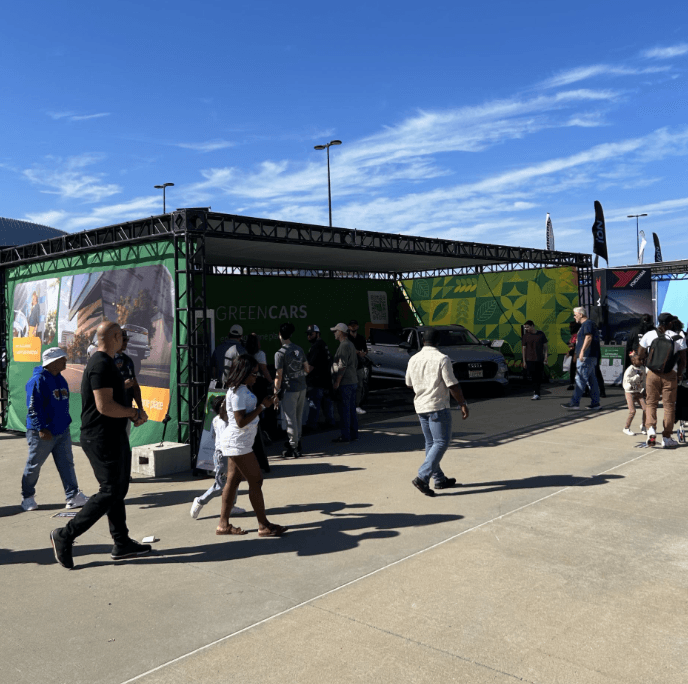 Vista amplia del stand de GreenCars con personas caminando junto a un vehículo estacionado.