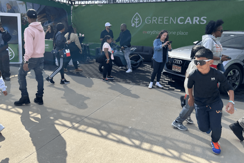 Varias personas pasan junto a un stand con un SUV plateado estacionado al lado.