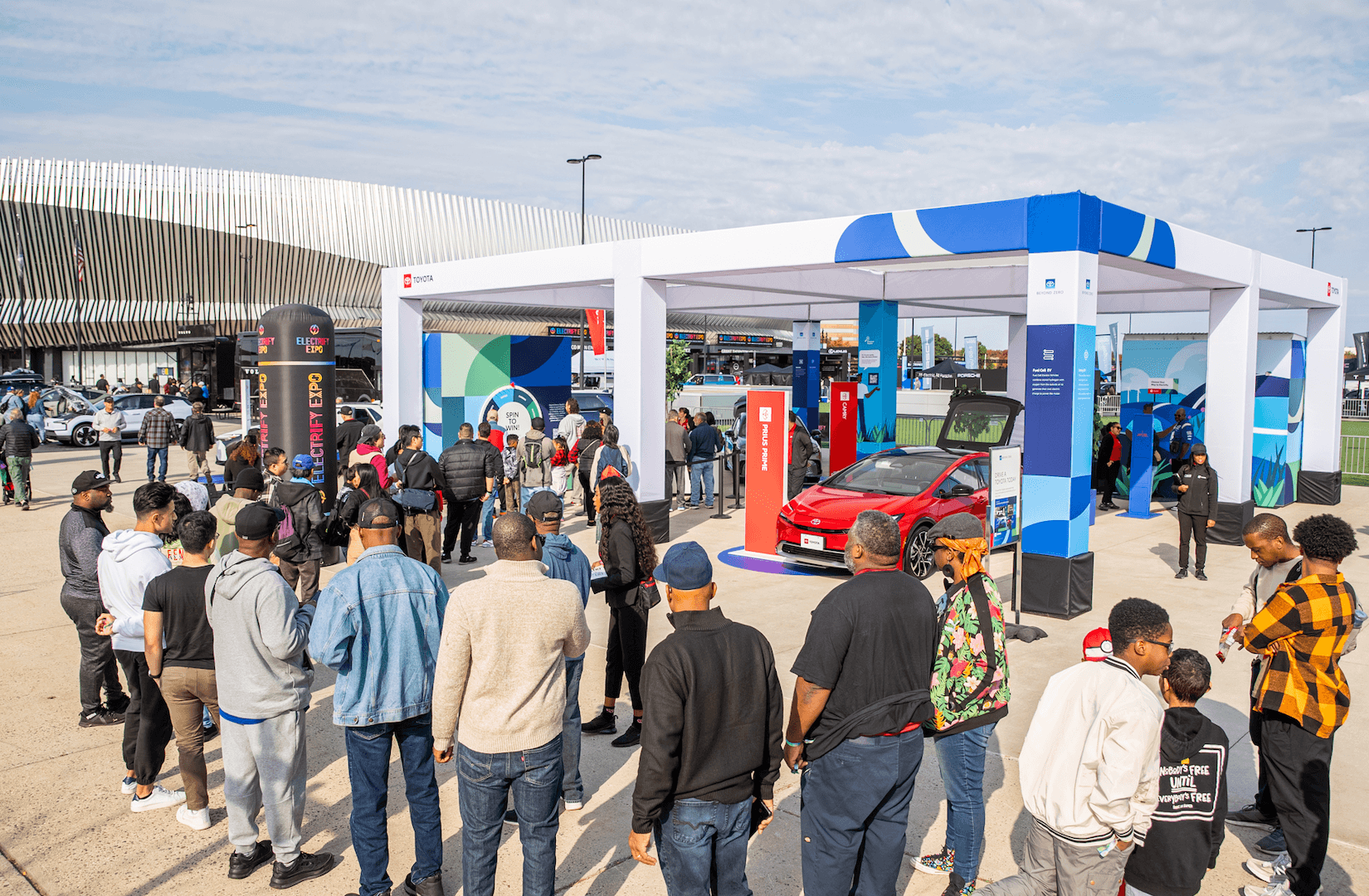 Una multitud se reúne alrededor de un auto híbrido rojo bajo un puesto de Electrify Expo.