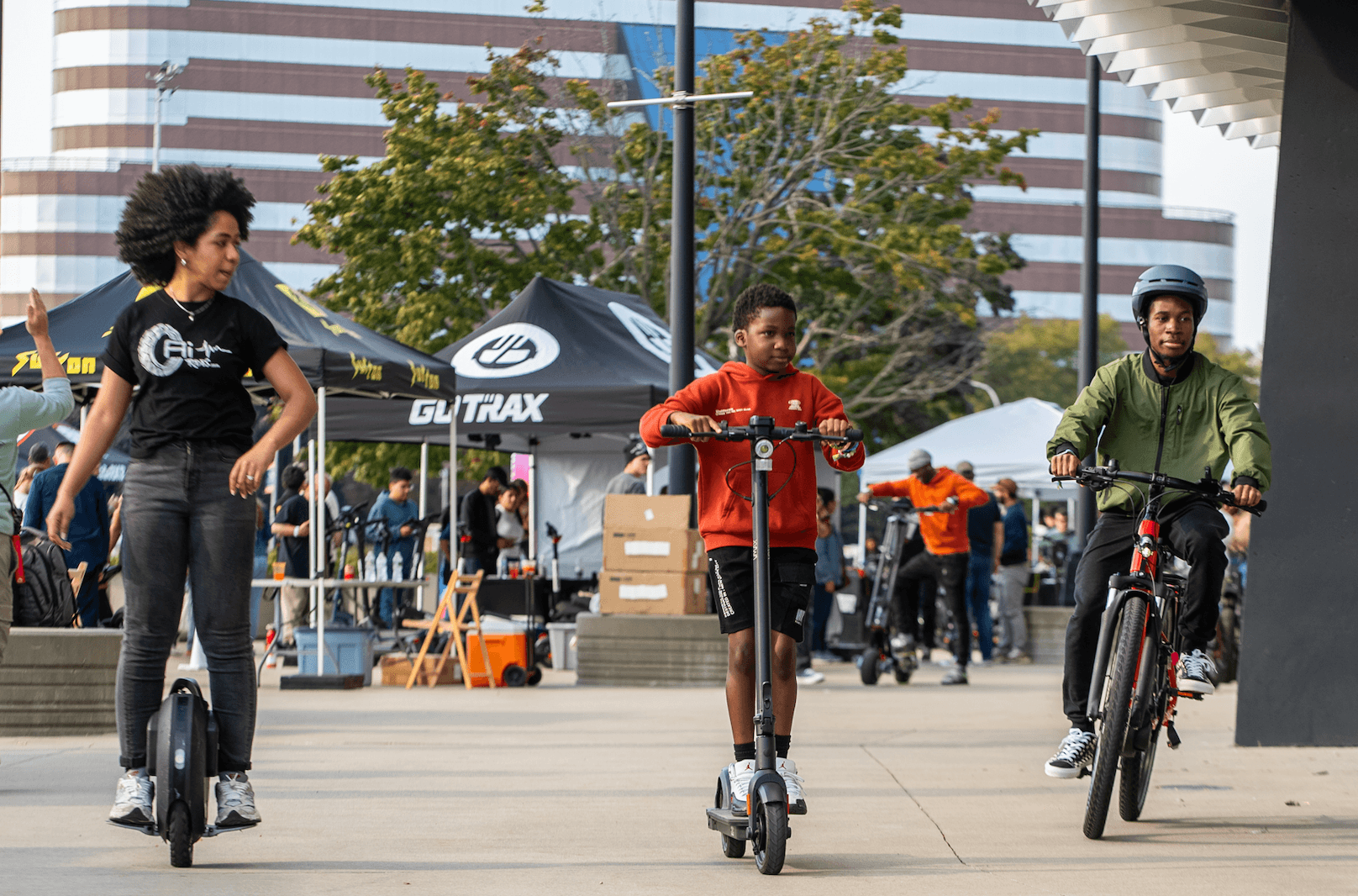 Tres personas andan en scooters y una bicicleta en una expo al aire libre.