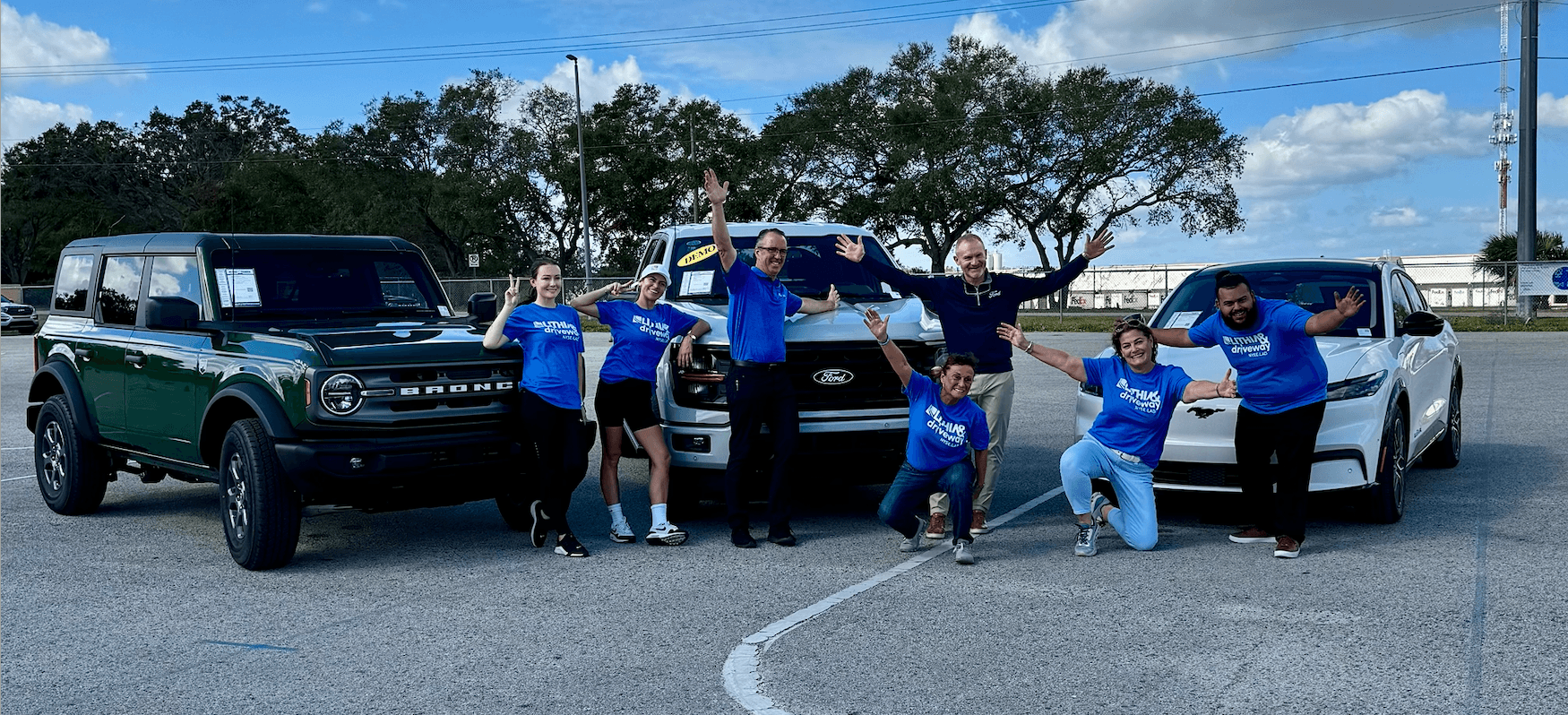Six people pose between three parked vehicles in a lot, waving at the camera.