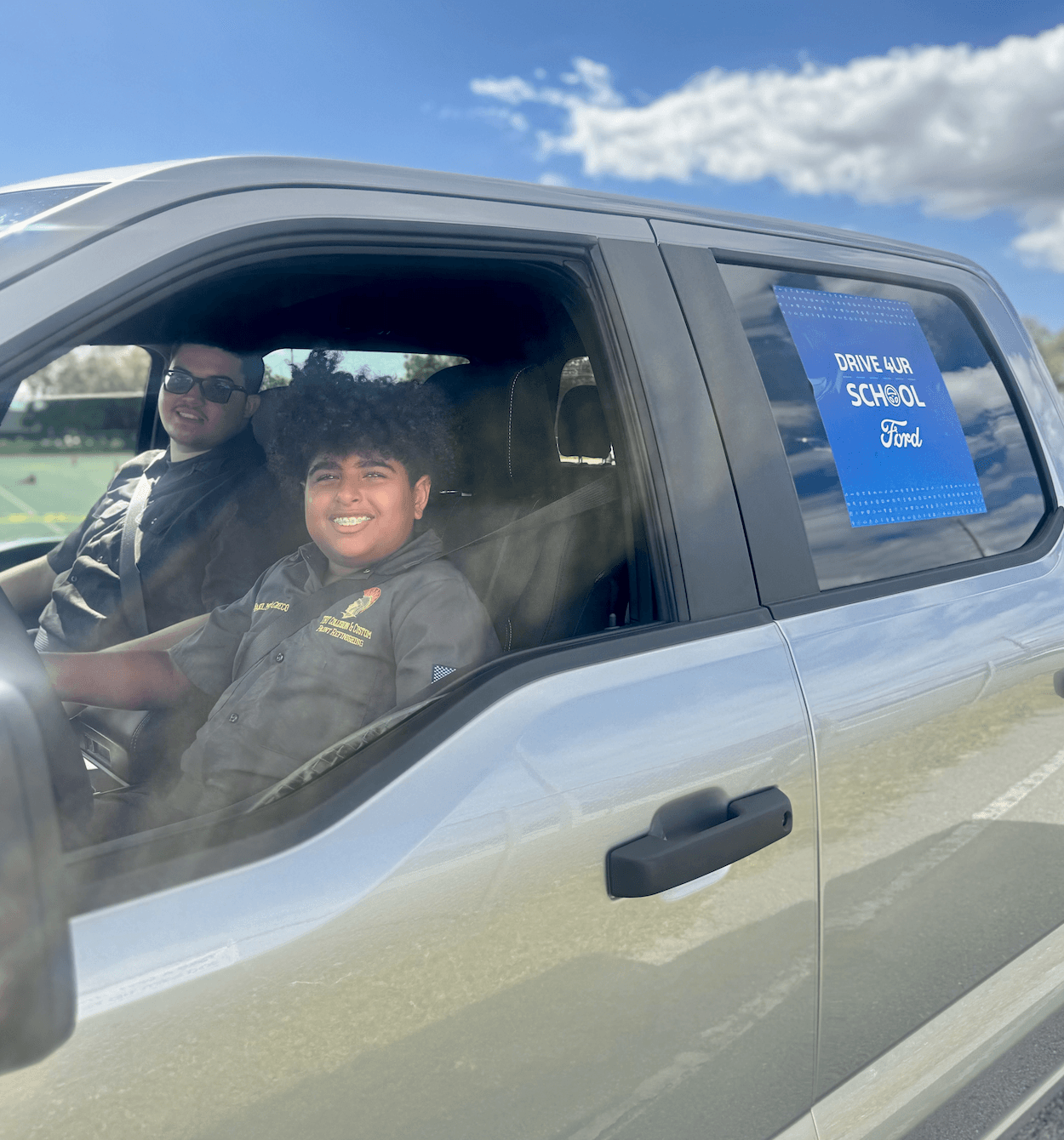 Side view of a silver Ford truck with two people inside and a Drive 4 UR School sign in the rear window.