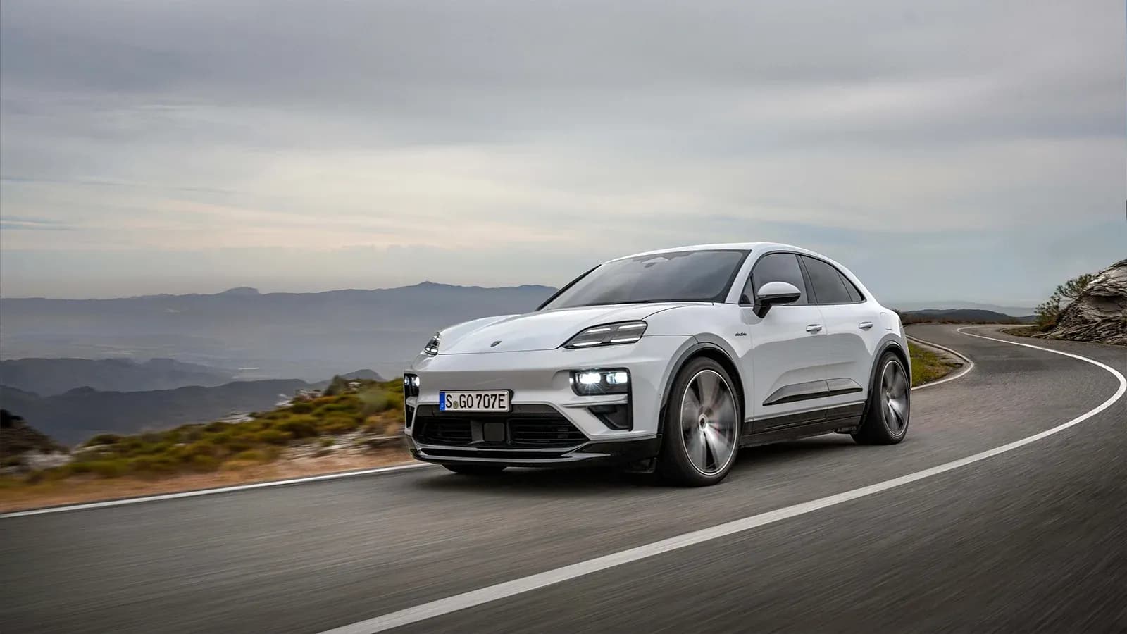 A white Porsche Macan EV drives on a winding mountain road.