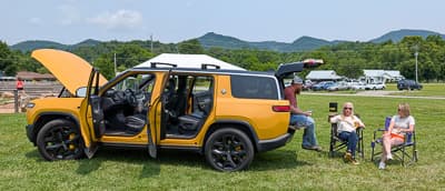 Vista lateral de una SUV eléctrica amarilla con ambas puertas del lado derecho abiertas y la cajuela trasera levantada en un evento al aire libre.