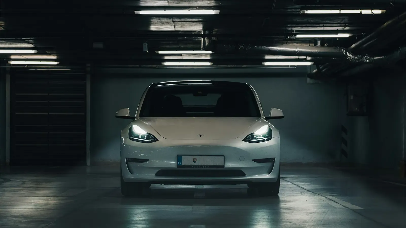 A white tesla model s in a dark parking garage with the headlights on