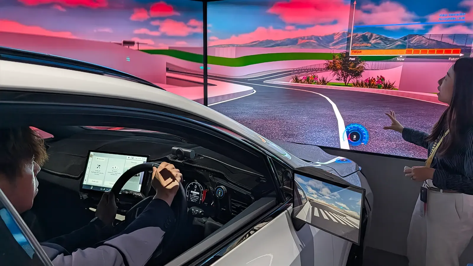 A person in a car uses a large screen while another person points at a road simulation display.
