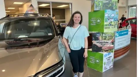 Front three-quarter view of a tan Subaru SUV beside a person standing next to Earth Day display boxes.