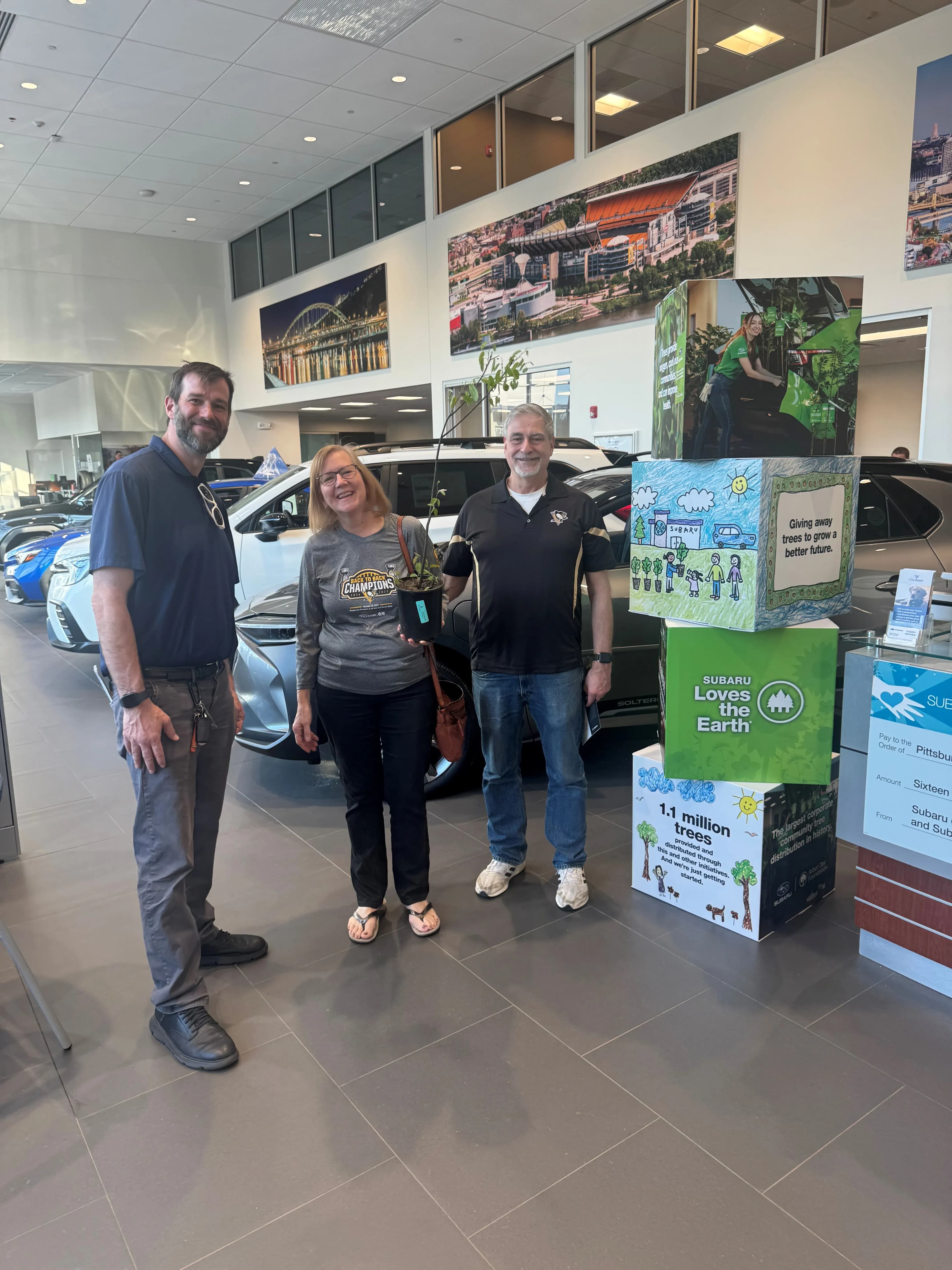 Three people stand in a Subaru showroom beside Earth Day displays and a potted sapling.
