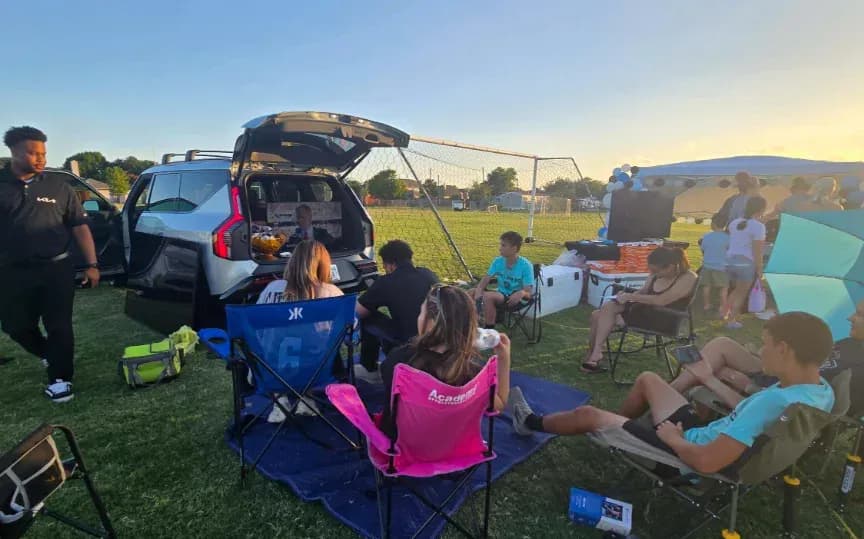 Vista trasera de una Kia EV9 con la compuerta trasera abierta mientras varias personas se sientan cerca en un evento en un campo.