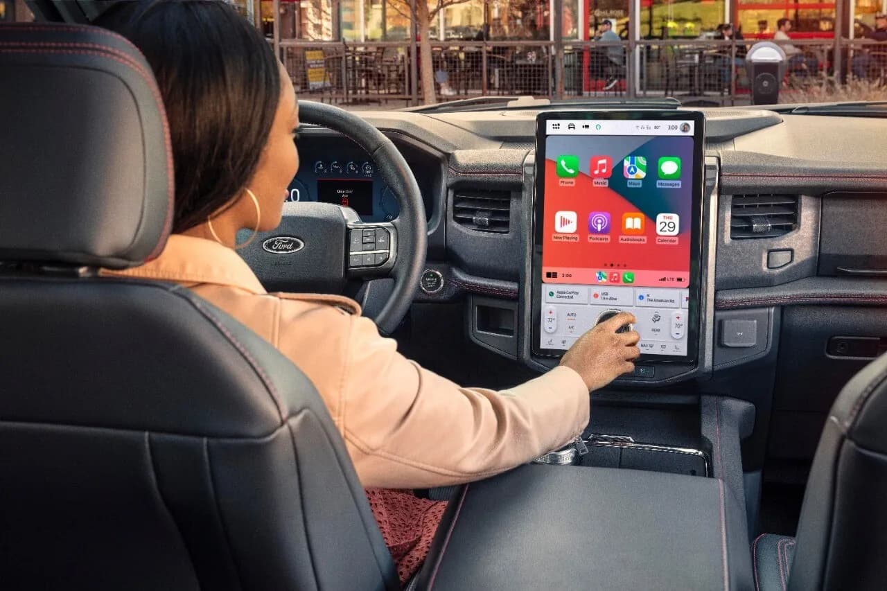 Driver seated in a Ford cabin using the center touchscreen with Apple CarPlay and Maps icons.