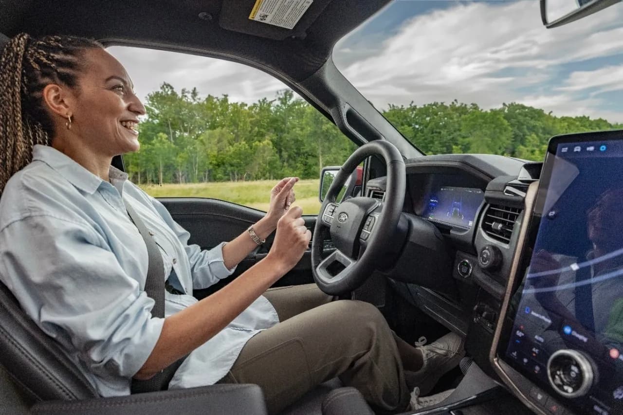 Interior view of a person sitting in the driver's seat with hands raised near the steering wheel and a large center screen.